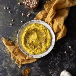 Bowl with turmeric hummus, yellow napkin under the bowl, small wooden bowl with garbanzo beans, small wooden bowl with turmeric powder and a wooden spoon on the side with turmeric on and off the spoon, garlic head and a lemon