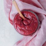 Pink towel wrapped around a glass jar filled with rhubarb strawberry butter and a wooden spoon inside the jar.