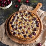 Cherry Clafoutis on a round wooden board, lying on top of a brown paper. Fresh cherries scattered around. And a bowl or fresh cherries on the side.