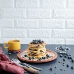 Stack of pancakes on a plate topped with shaved chocolate and fresh blueberries. Yellow cup of coffee on the left side, small glass bowl full with fresh blueberries on the right side with blueberries and chocolate scattered on the table. Fork and knife by the side of the plate with a pink napkin next to them.
