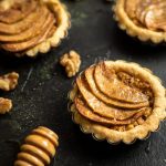 Three mini Apple Walnut Tartlets, fresh walnut pieces scattered around. Wooden honey dipper in the bottom left corner.