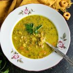 Bowl of Split Pea Vegetable Soup with a spoon in it. Fresh parsley at the bottom left corner. Yellow napkin at the top side of the bowl. Wooden bowl with crispy round pretzels, some scattered around the bowl of soup.