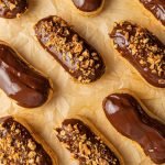 Eclairs arranged diagonally on a parchment paper. All of them are dipped in chocolate and some have also chopped nuts on top.