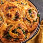 A round baking dish with baked rolls filled with cheese, sun-dried tomatoes, and cheese. Yellow napkin around the dish. Spinach leaves on the napkin and next to the dish.