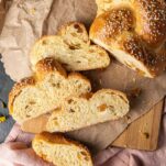 Challah bread with a four slices on a brown paper on top of a wooden board. Pink napkin underneath. Few raisins on the side.