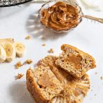 Banana dulce de leche Muffins on a black round wire tray. On the left side one banana chopped into slices. On the right side white napkin laying next to the tray. Small glass bowl with dulce de leche and a spoon. One muffin cut in half in the front with crumbs around it.