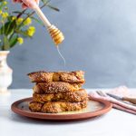 A plate with four pieces of Challah French Toast stacked one on top of each other. A hand holding honey spoon, drizzling honey over the toast. Napkin next to the plate with fork and knife on it. A vase on the back left side with flowers, green and yellow.