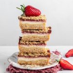 Four Strawberry Cream Cheese Bars stacked up one on top of each other with a strawberry on top. Sitting on a plate and a pink ruffed edges napkin under the plate. Sliced strawberry on the side, mint leaves and crumbs.