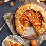 Apricot Ricotta Galette on a plate and a slice cut out. Small plate with the cut out slice and a fork next to the galette. Baking paper under the plate. Fresh peaches scattered around. Small glass ramekin with honey and honey spoon.