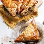 A plate with Phyllo dough turnover. Behind another plate with four turnovers and pieces of walnuts on a plate. The plate is sitting on a cutting board partially covered with yellow napkin. Small whole pumpkin on the back.