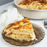 Two slices of Banitsa one on top of the other on a dark glass plate. Two forks next to the plate. Round baking dish with Banitsa in it on the background. White napkin on the side next to the plate.