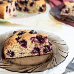 Slice of a Blueberry yogurt cake on a dark glass plate. A fork next to it. In the background a cake with a slice on the side and a dark pink napkin under the plate.