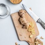 Wooden cutting board with a ginger and slices from it. A knife on the side. Ginger shavings around the board and a potato peeler. A measuring cup with a brown sugar in it. A glass with water next to the cutting board.