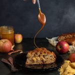 A slice of apple cake on a plate and a hand drizzling caramel over the slice. Two forks next to the plate with a few apple peels in front. Whole apples on both sides in the back of the plate. with a jar of caramel and more cake. The corner of a yellow napkin peeking out form the right side next to the plate.