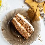 A dark glass plate with a slice of Carrot Cake and a fork next to it. Small ramekin with chopped walnuts and a yellow napkin underneath.