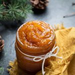 Pumpkin jam in a jar with a white string around it tied into a bow tie. The jar is sitting on a yellow napkin. Pine cones in the background with pine branches.