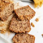 Banana walnut bread laying on a parchment paper with three slices cut out. Pieces of walnuts scattered around. A knife on the side with a yellow napkin next to it.
