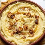 A dish with a brown butter mashed potatoes, roasted pieces of garlic, and herbs on top. Yellow napkin underneath the plate and the end of a utensil visible on it. Fresh garlic clove on the back of the plate.