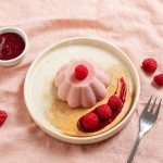 Panna Cotta on a plate with raspberry coulis and fresh raspberries. Fork on the side and ramekin with raspberry coulis. Pink tablecloth underneath.